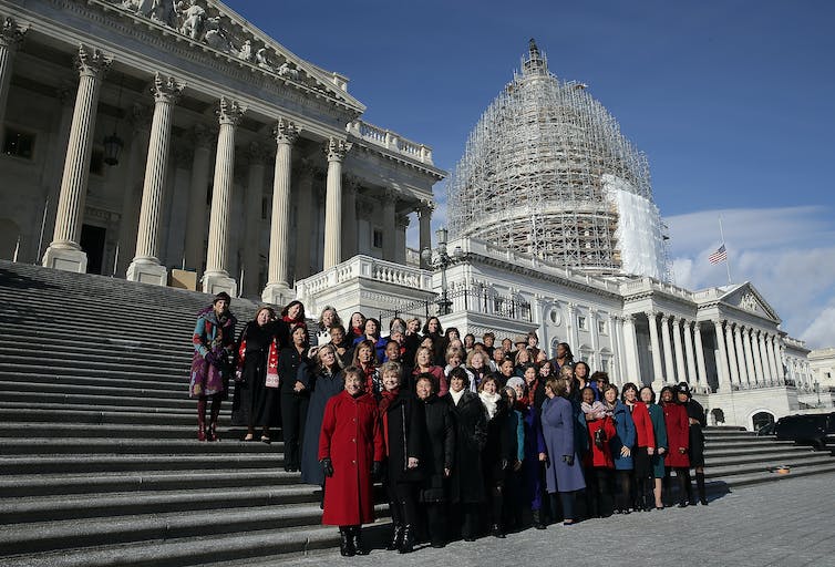 A large group of women wearing jackets stand on a flight of outdoor steps, with two white buildings with pillars behind them.